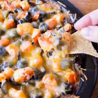 a hand with a tortilla chip digging into a black plate covered with a dip consisting of refried beans, olives, tomatoes, ground beef, and melted cheese.
