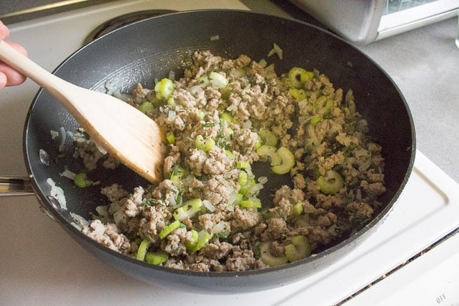 Cut up celery, onions, fresh herbs, and sausage being stirred by a wooden spoon in a large black pan on a stovetop.