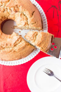 A circular light brown pound cake on a white stand on top of a red tablecloth with one slice being taken out of it. A white small plate with a fork are in the bottom left corner.