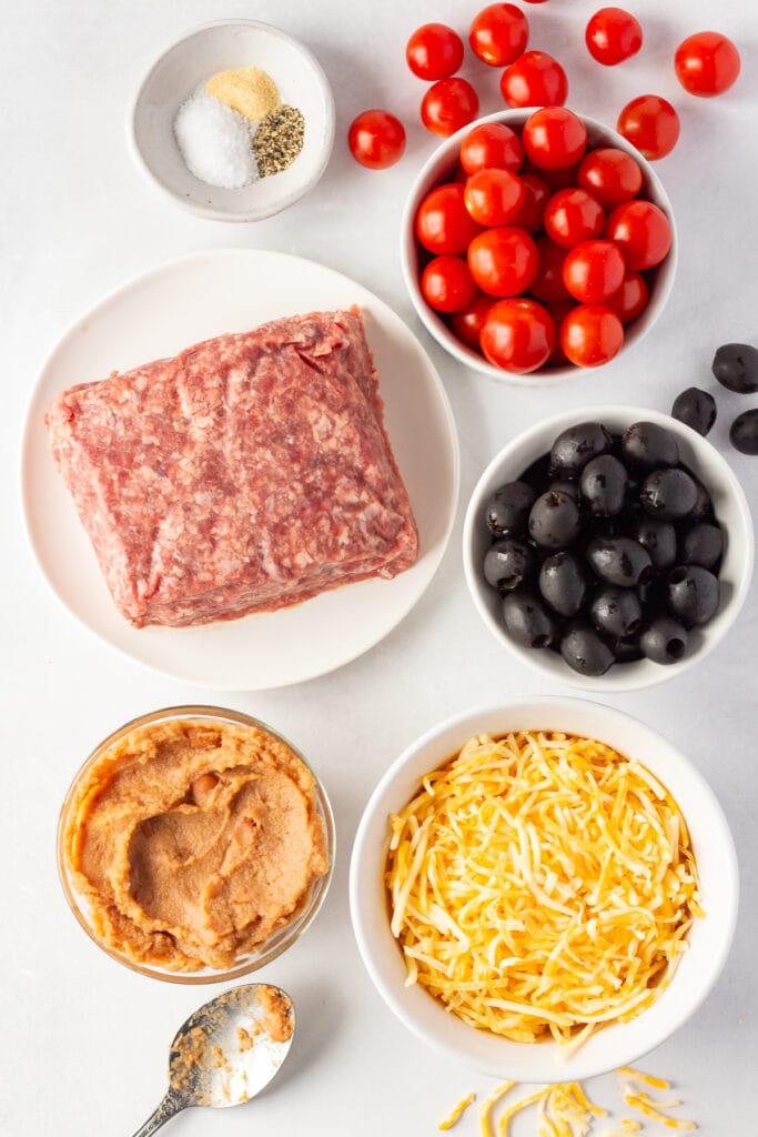 Top down shot of ingredients for ground beef bean dip on a white background, including plates and bowls with raw ground beef, black olives, red cherry tomatoes, refried beans, spices, and shredded cheese.