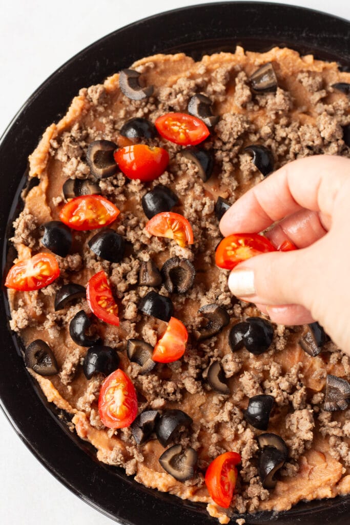 A hand sprinkling cut cherry tomatoes onto a black plate that has refried beans, ground beef, and cut black olives already on it.