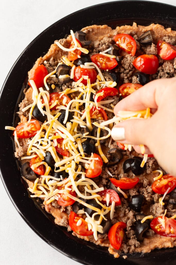 A hand sprinkling shredded cheese on top of layers of refried beans, ground beef, and cut red cherry tomatoes and black olives.
