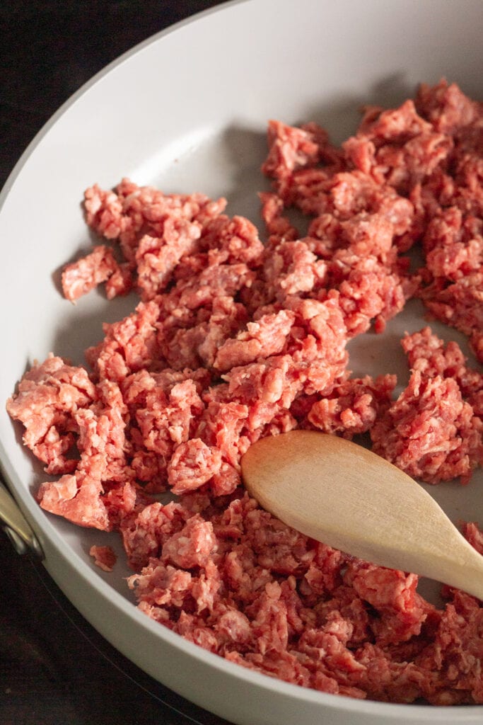 Raw ground beef being cooked in a gray large pan on a stove top. A wooden spoon rests in the pan as well.