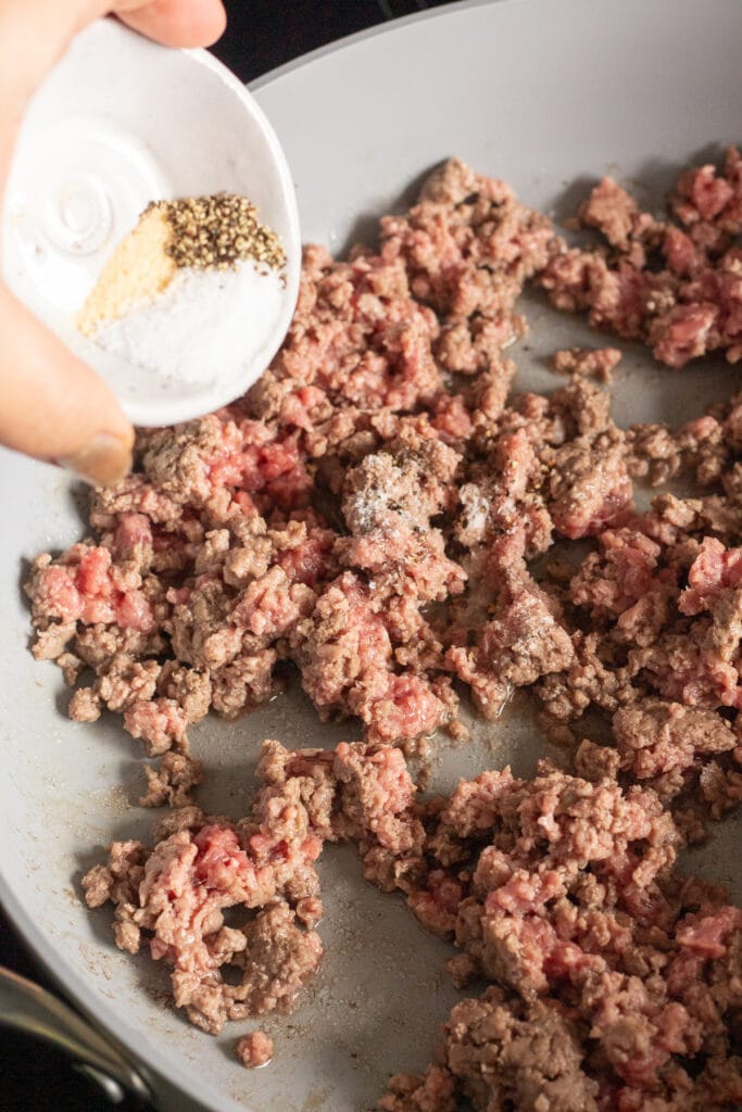 A hand pouring spices from a small white bowl into a large gray pan that's cooking ground beef.