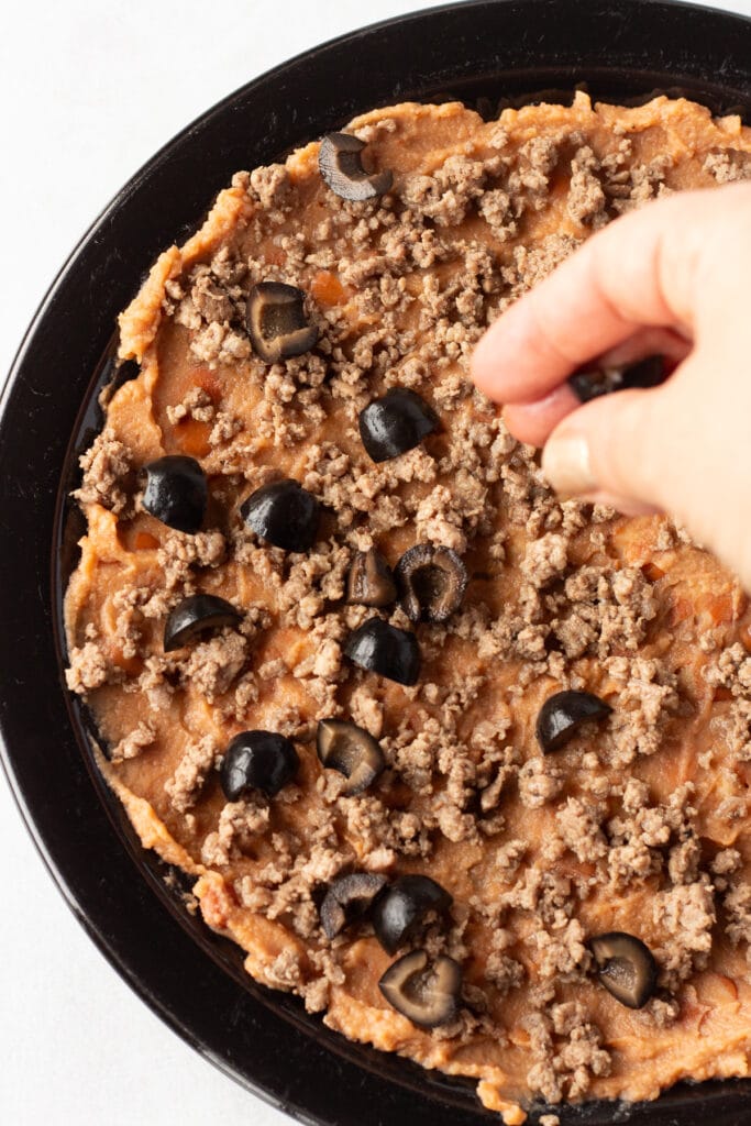 A hand sprinkling cut olives onto a black plate with refried beans and ground beef already on it.