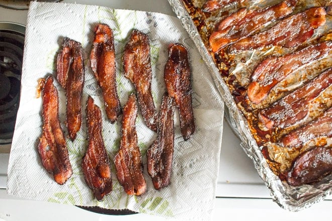 Top down shot of cooked bacon on a plate lines with paper towels. A sheet pan with more cooked bacon on it is to the right of the plate.