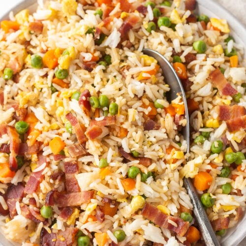 Top down shot of a large gray bowl with bacon fried rice in it, and a large silver serving spoon resting in it to the right.