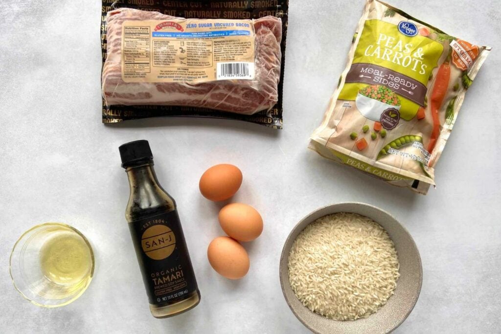 Top down shot of ingredients for bacon rice on a light gray background, including eggs, tamari, bacon, frozen veggies, and white rice.