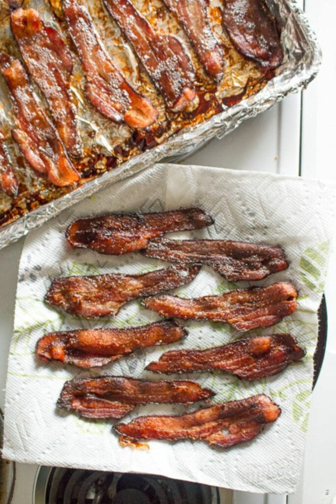 Top down shot of cooked bacon on a paper towel lined plate with a sheet pan showing more cooking bacon next to it.