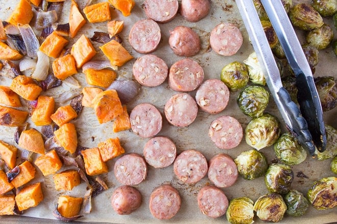 Horizontal image of a sheet pan with roasted sweet potatoes, red onions, Brussels sprouts, and sliced chicken apple sausage with a pair of tongs lying on top.