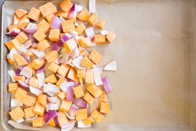 Top down shot of uncooked and cut up sweet potato and red onion on the left half of a sheet pan lined with brown parchment paper.