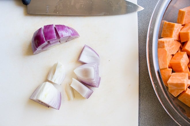 Top down shot of red onion being sliced into chunks on a white cutting board. A large bowl with cut up sweet potato is to the right of the board.