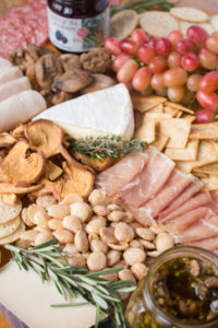 A close up of a cheese board with meats, cheese, fruit, nuts, jam, and garnishes on it.