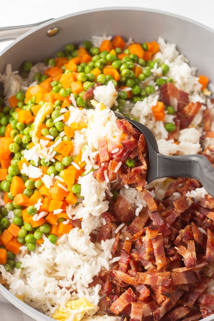 A black spatula combining peas, carrots, sliced bacon, egg, and white rice all together.