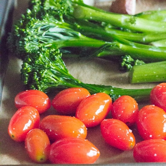 Close up of cherry tomatoes and broccolini on a sheet pan lined with brown parchment paper.