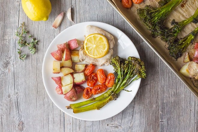 Top down shot of chicken, potatoes, roasted tomatoes, and broccolini on a white plate on top of a gray wooden background.
