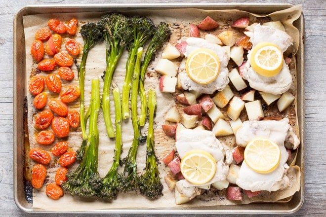 Top down shot of roasted tomatoes, broccolini, cut red potatoes and chicken thighs topped with a lemon slice on a sheet pan.