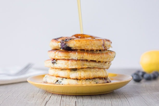 Maple Syrup being poured over a stack of Gluten Free Gingery Lemon Blueberry Pancakes on a yellow plate