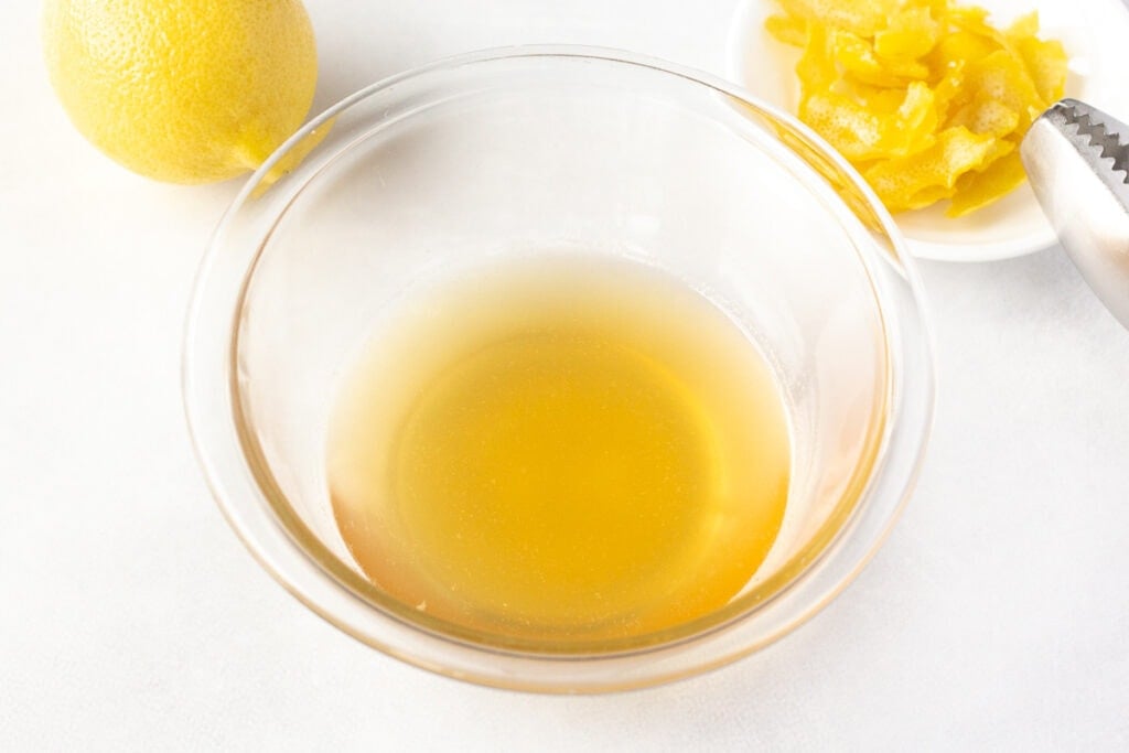 a clear bowl with lemon syrup in it, with a lemon and lemon skin in the background.