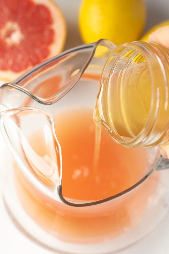 pouring lemon simple syrup into a glass pitcher with grapefruit juice in it.
