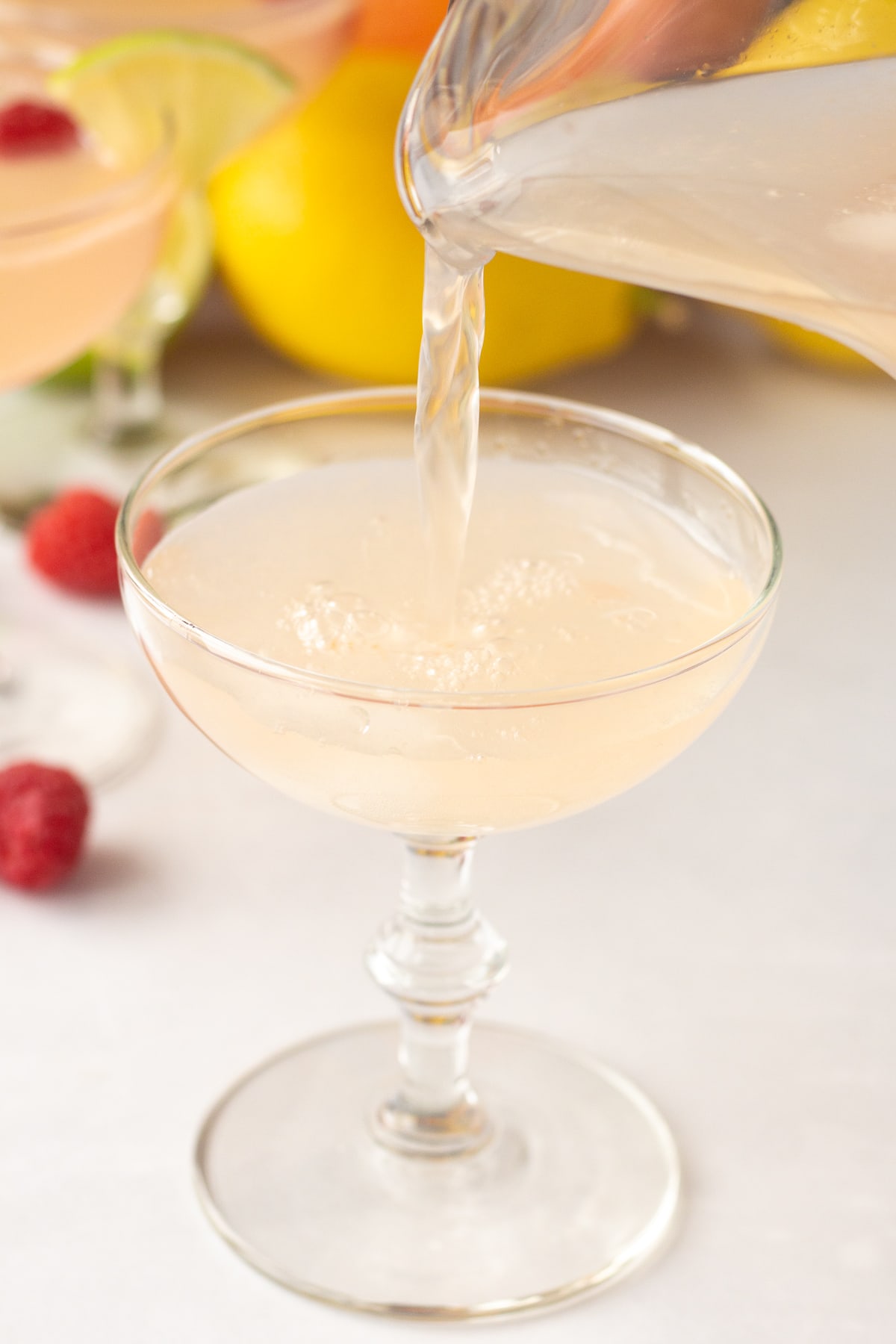 pouring a pink citrus mocktail from a glass pitcher into a champagne glass.