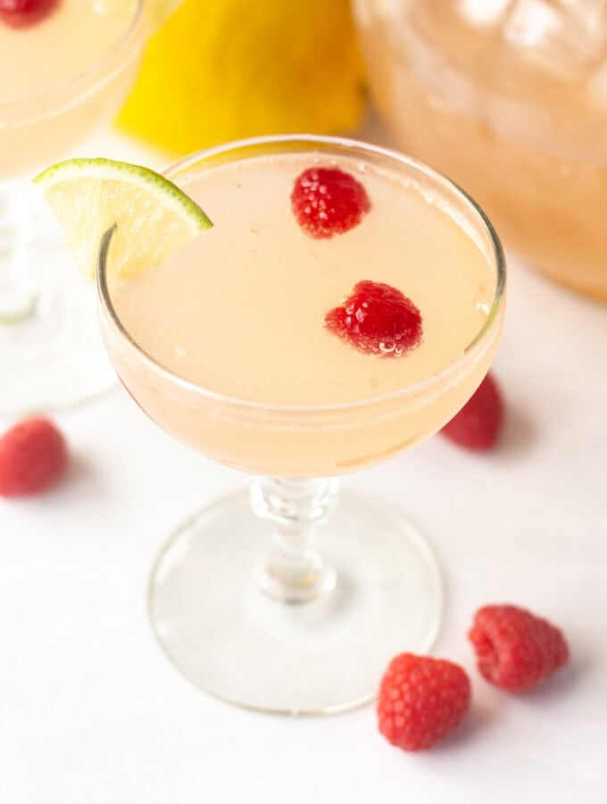an angled shot of a pink mocktail in a low champagne glass with raspberries and a lime wedge in it.