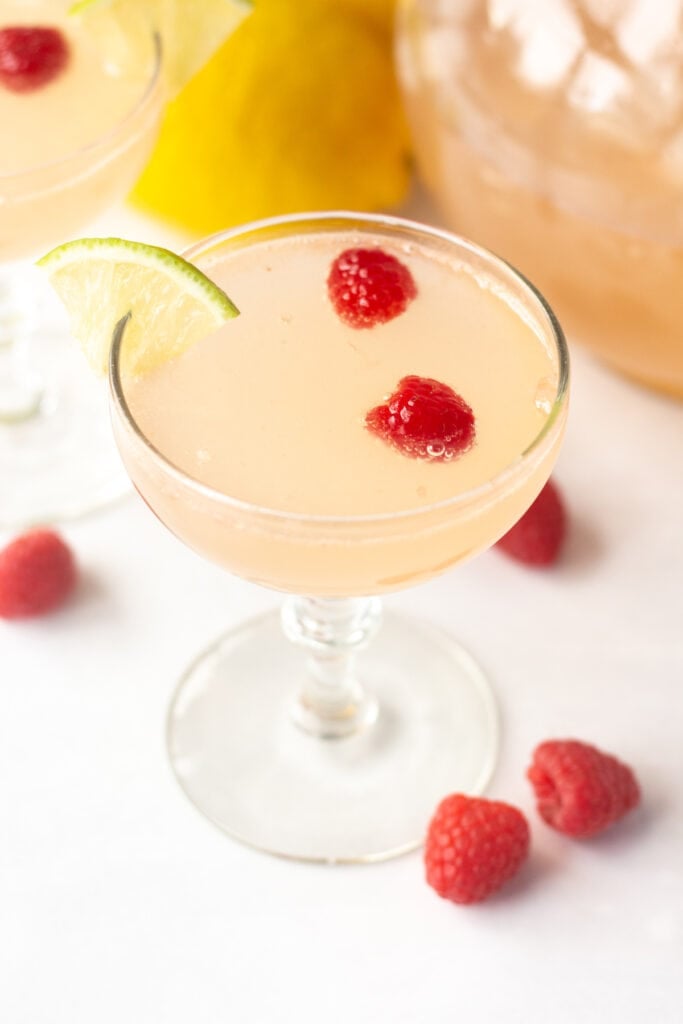 an angled shot of a pink mocktail in a low champagne glass with raspberries and a lime wedge in it.