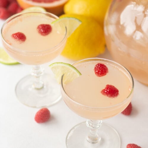 two fancy glasses with pink mocktails in them, garnished with raspberries and lime wedges.
