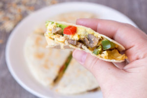 A hand holding a quesadilla with eggs, mushrooms, and red and green peppers in it.