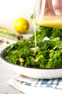 A hand pouring dressing from a mason jar onto spring salad in a low white bowl with asparagus, lemon, and lettuce in the background.