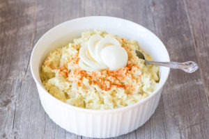 A large white bowl with gluten free potato salad in it and a spoon on the right side. It's topped with a sliced hard boiled egg and some paprika.