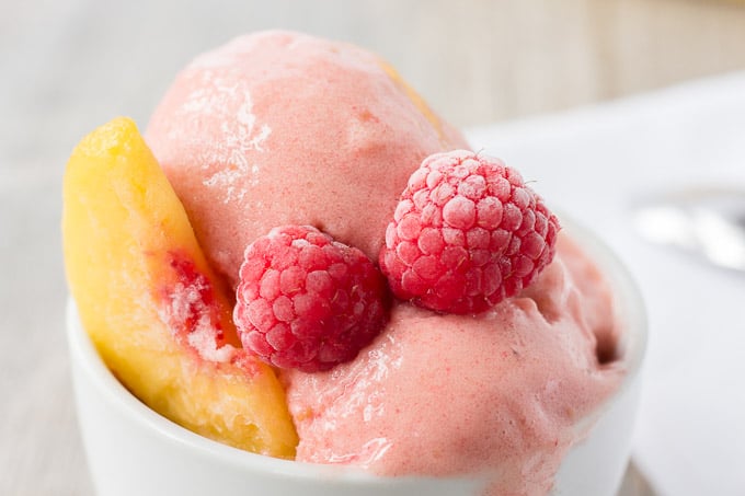 close up of raspberry peach nice cream slightly melting in a white bowl with frozen fruit also in the bowl.
