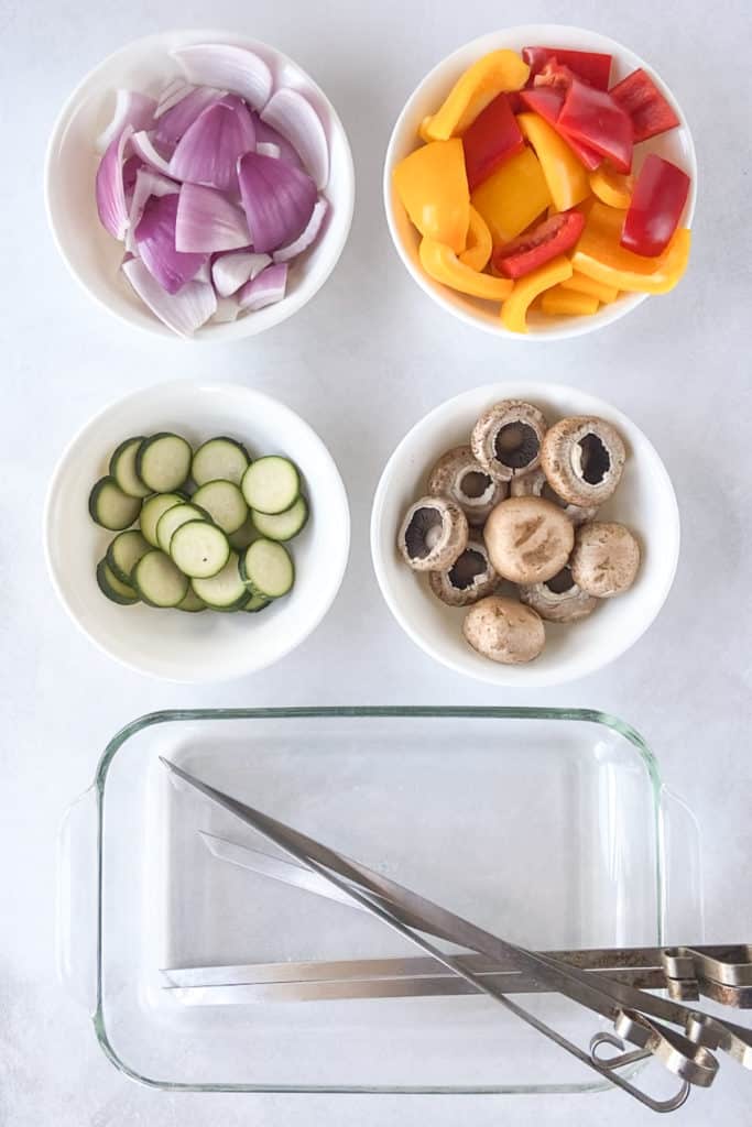 Cut up red onions, bell peppers, zucchini, and mushrooms in white bowls next to a clear glass dish with metal skewers in it.