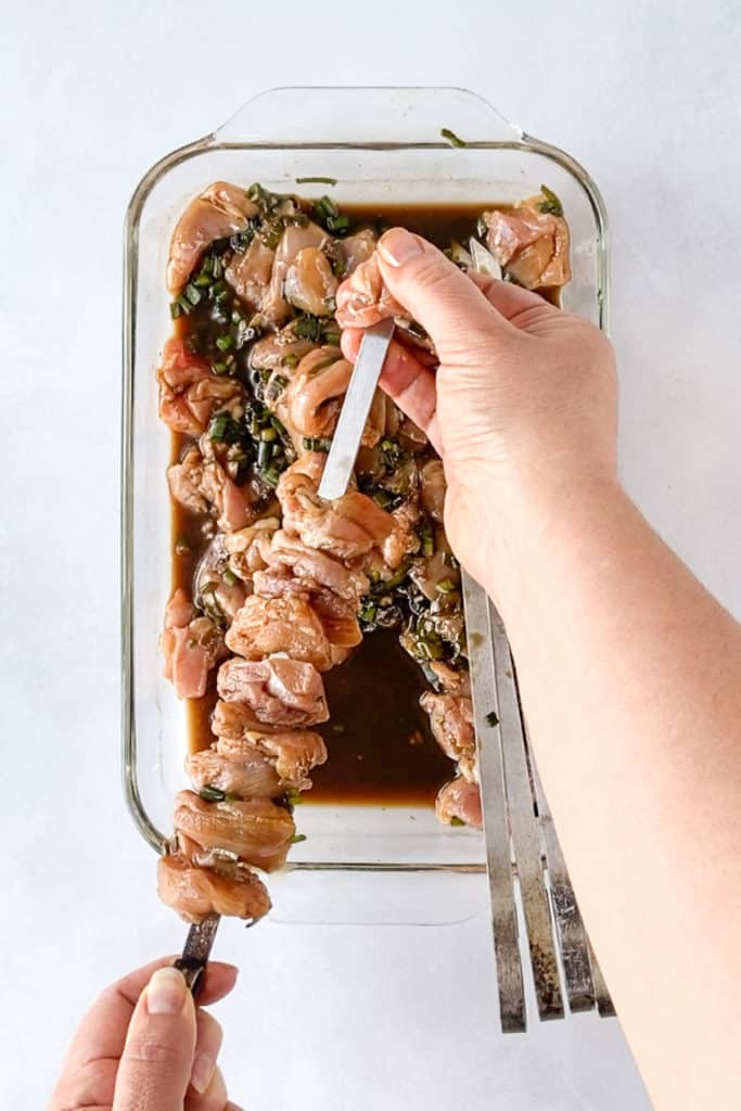 Two hands putting marinated chicken pieces on a metal skewer.
