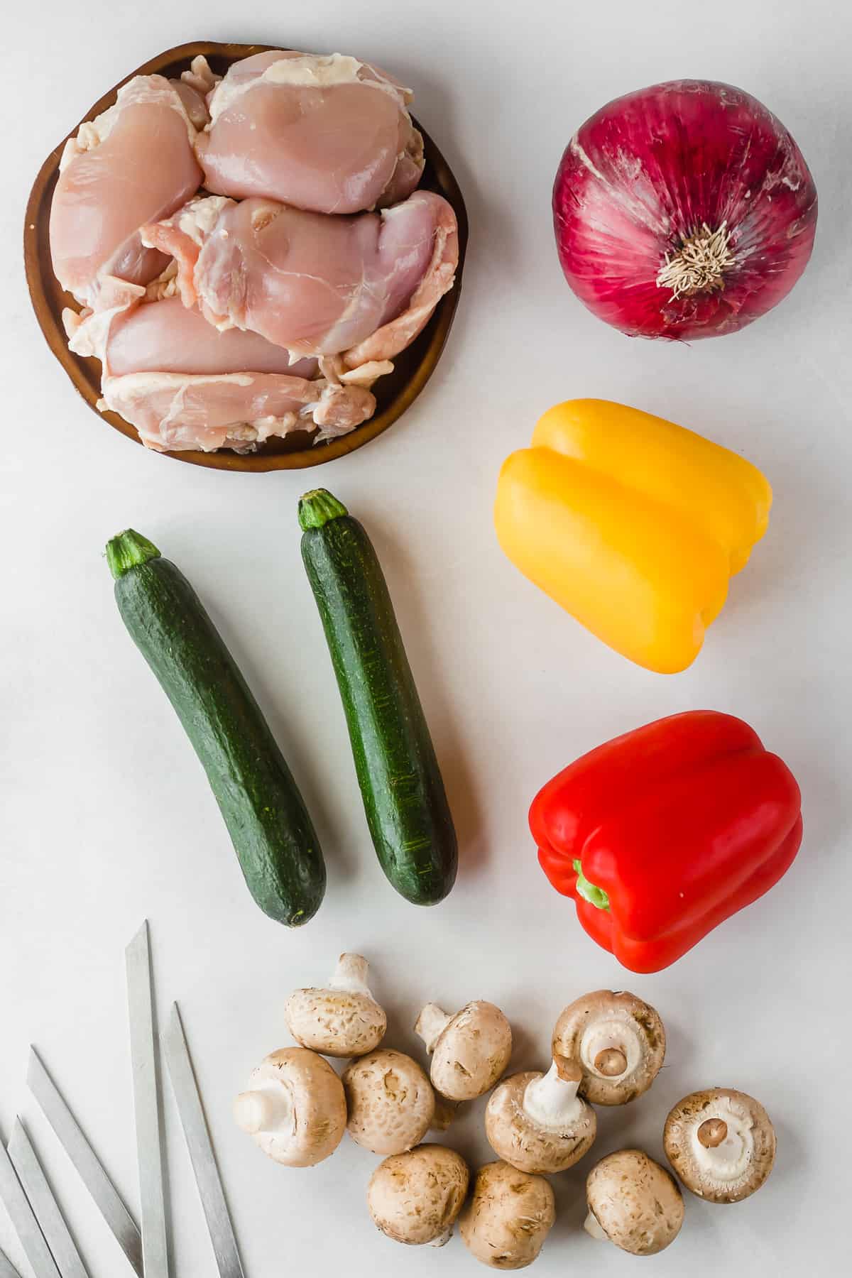 Top down shot of veggies, raw chicken thighs, and metal skewers on a white background.