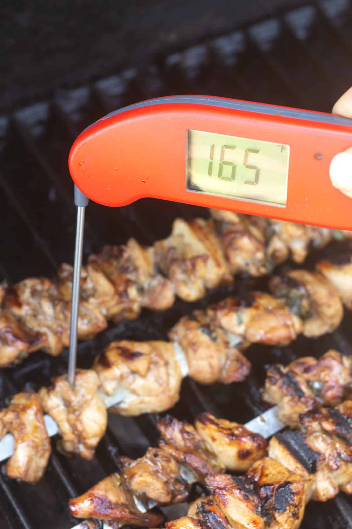 A hand measuring the internal temp of chicken kabobs on the grill.