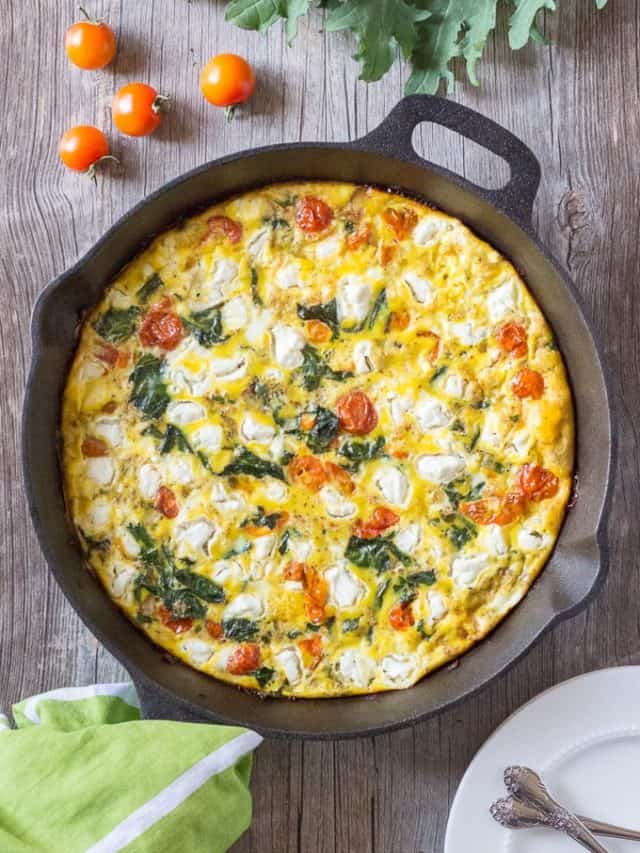 Top down shot of a frittata with goat cheese, roasted tomatoes, and kale in a black cast iron pan. It rests on a gray wooden surface with several fresh cherry tomatoes and kale at the top of the frame and a green and white towel and white plate at the bottom of the frame.