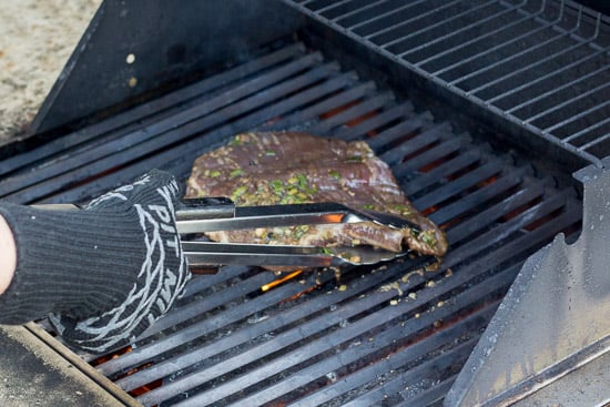 A gluten free flank steak being placed on a hot grill metal tongs.