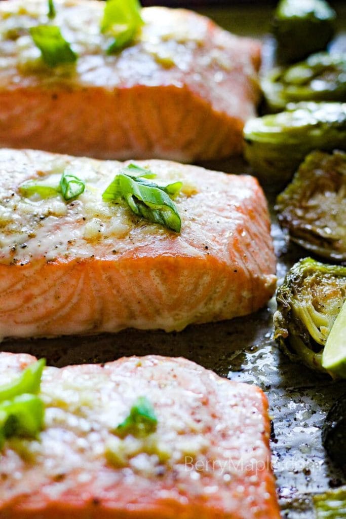 Close up of cooked salmon filets on a sheet pan with roasted Brussels sprouts next to them and sliced green onions on top.
