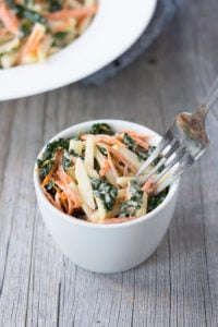 Close up of a small white bowl with coleslaw made from kale, kohlrabi, and sliced carrots in it. A fork is coming in from the right side and a larger white bowl with the same coleslaw is in the background.