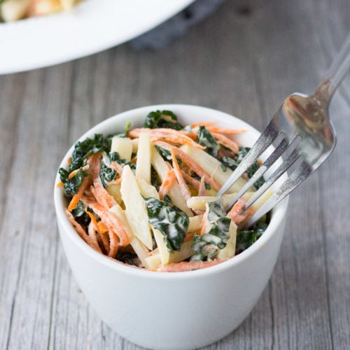 Close up of a small white bowl with a coleslaw made from shredded carrots, kale, and kohlrabi and a fork sticking into it.