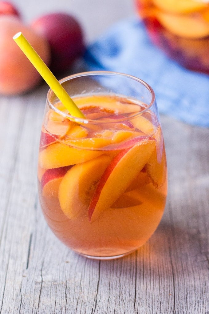 stemless glass of peach rose sangria with fruit in left background and clear pitcher with sangria on a blue towel in right background