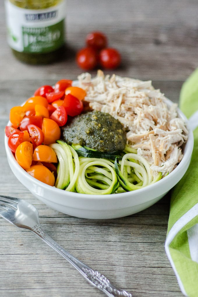 white bowl with pesto, chicken, zoodles, and cut tomatoes on a gray background with a white and green towel on the right, silver fork on the left, and pesto container and 3 cherry tomatoes in the background