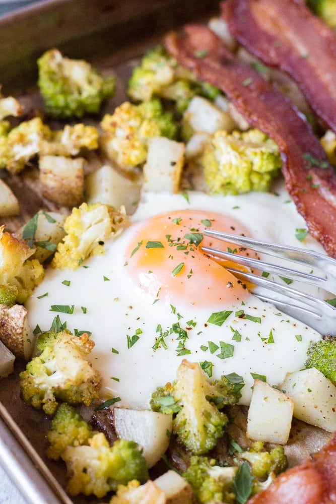 Close up shot of a silver fork cutting into a cooked egg on a sheet pan with potatoes, romanesco, and bacon.