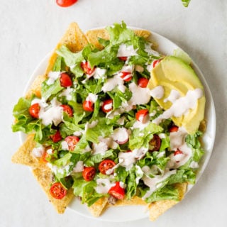 white plate with nachos, lettuce, cut cherry tomatoes, and dressing on top, with slices of avacado on the upper right. On a white background with several tomatoes and some lettuce up top and two tortilla chips on bottom right