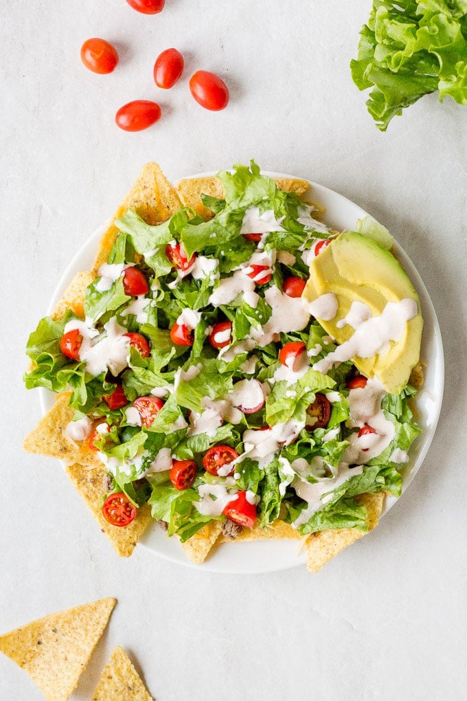 Top down shot of a white plate with nachos, lettuce, cut cherry tomatoes, and dressing on top, with slices of avacado on the upper right. On a white background with several tomatoes and some lettuce up top and two tortilla chips on bottom right.
