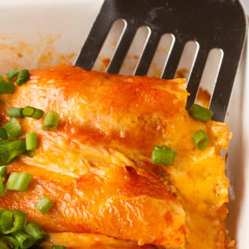 A metal spatula scooping enchiladas out of a white casserole dish.