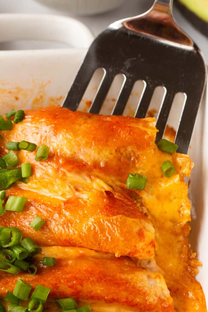 A metal spatula scooping enchiladas out of a white casserole dish.