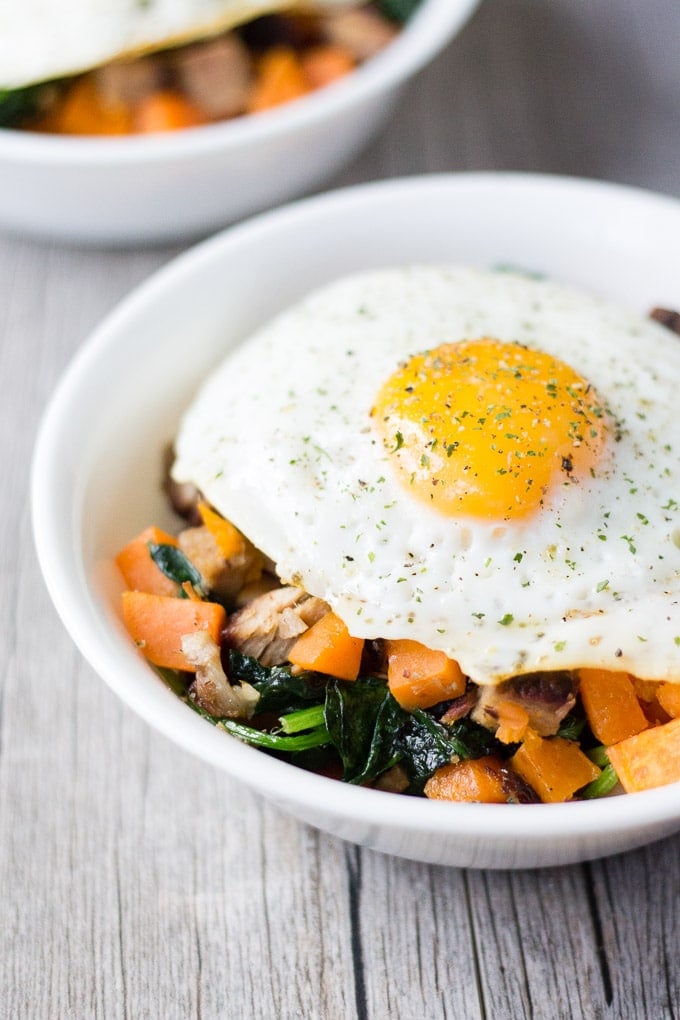 cut up sweet potatoes, pork, and spinach in a white bowl with an over-easy egg on top, on a gray background with another bowl of the same in the upper left