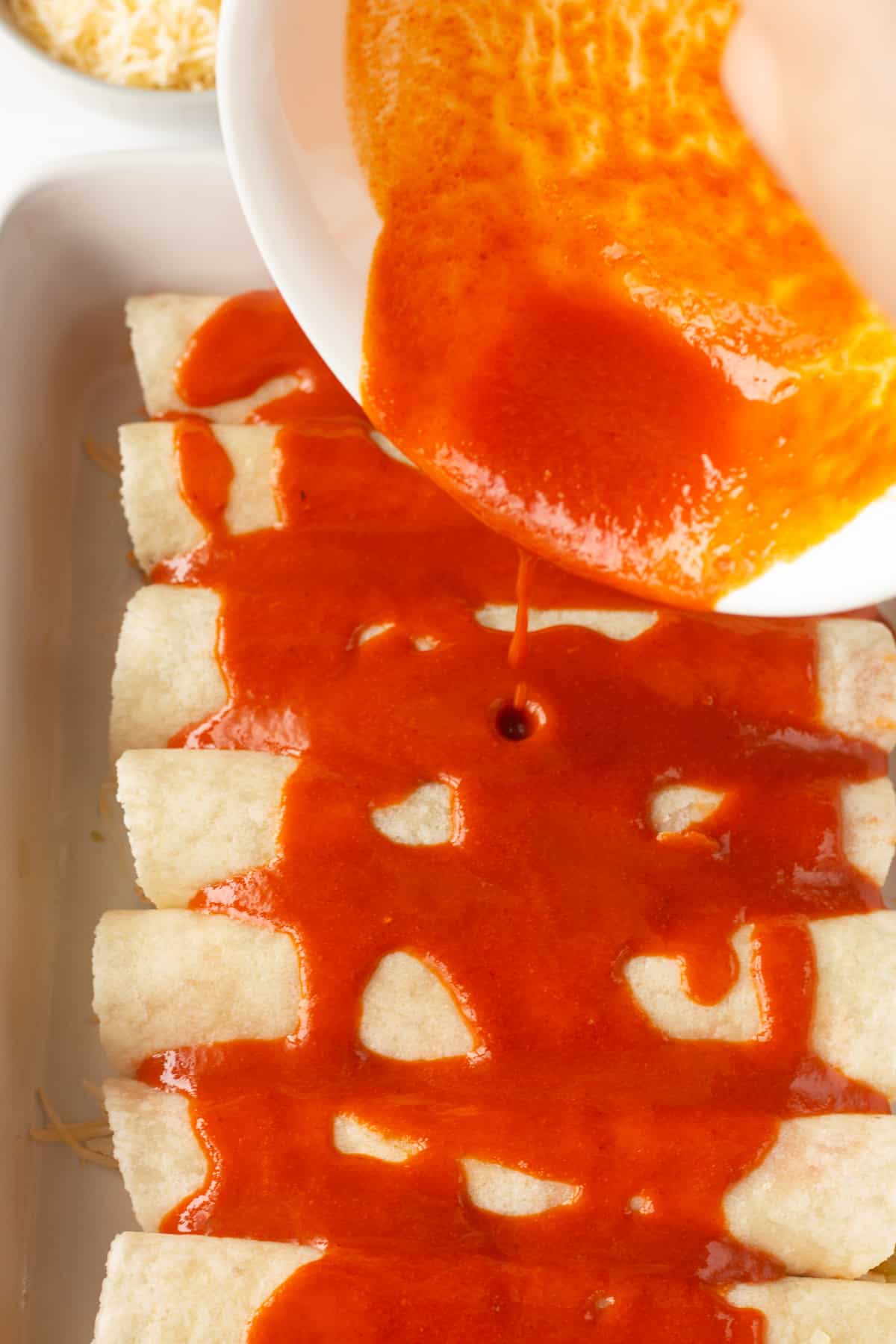 Pouring red enchilada sauce out of a white bowl onto rolled up uncooked enchiladas in a white casserole dish.